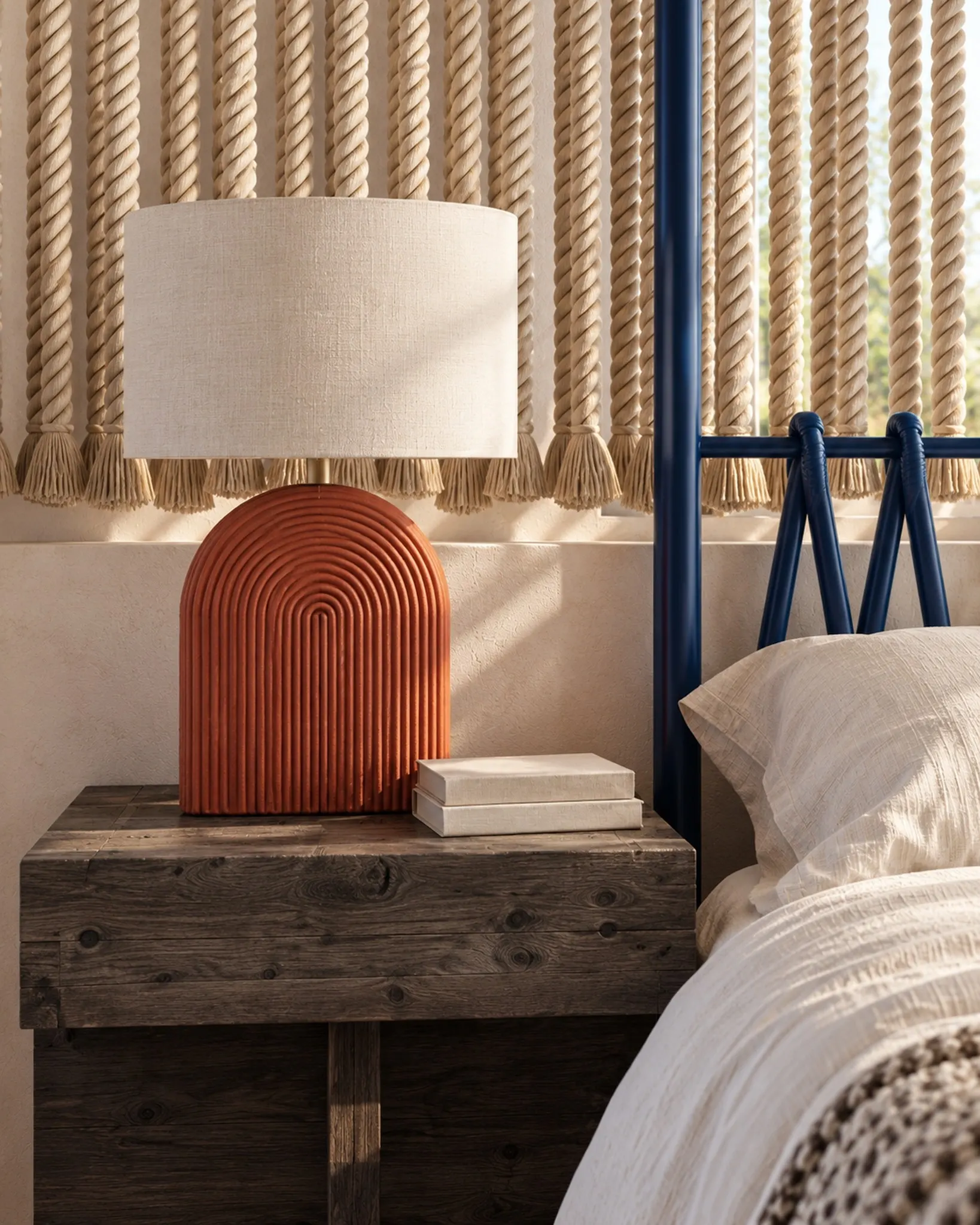 Bedside detail with a terracotta lamp on a rustic timber table, a rope screen and the blue bed frame.