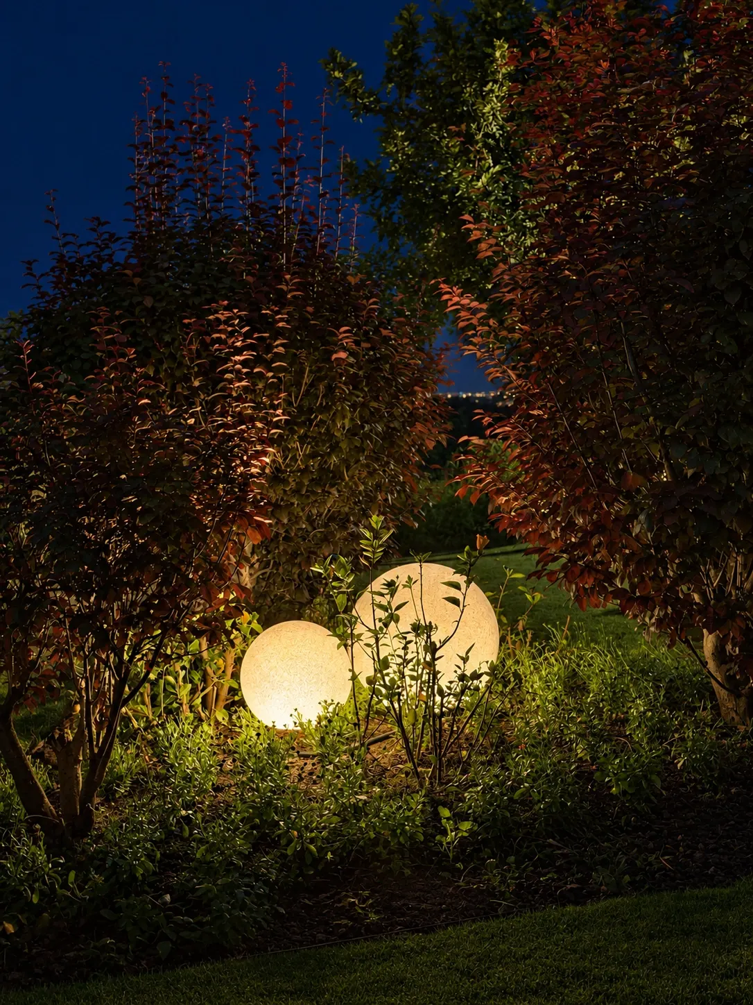 Two glowing spherical lights nestled among shrubs and low planting in an evening garden.